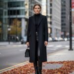 Žena v černém kabátě stojí na městské ulici s podzimními listy, městské pozadí rozmazané.